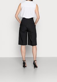 Žena stojící s rukama v kapsách, v bezramínkovém bílém topu, černých kalhotách do pasu a černých vysokých podpatcích.