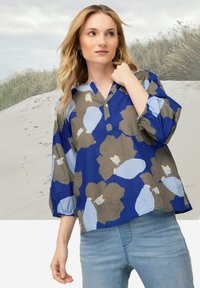 Blusa floreale blu con pattern marroni e blu chiaro, caratterizzata da una vestibilità comoda, scollatura a V e polsini elasticizzati. Abbinata a jeans denim.