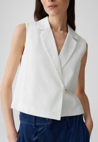 Blusa bianca senza maniche con colletto a scollatura, dotata di una chiusura a un solo bottone, tessuto testurizzato e design cropped. Abbinata a denim blu.