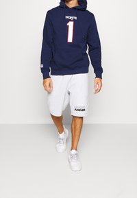 Sudadera azul marino con "PATRIOTS" y el número "1" en blanco, combinada con shorts grises que muestran el texto "UNDER ARMOUR", completada con zapatillas blancas.
