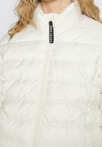 Kurtka puchowa w odcieniu ecru, z wysokim kołnierzem i poziomym pikowaniem. Zawiera zamek błyskawiczny z logo na suede.