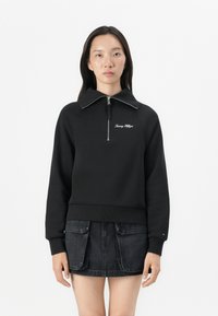 Black sweatshirt with a high collar and zip, featuring white "Tommy Hilfiger" embroidery on the chest. Smooth material with a fitted design.