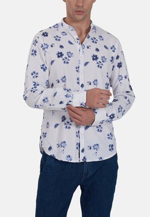 Uomo che indossa una camicia bianca con stampa floreale blu e jeans blu scuro, mentre sistema il polsino sinistro con la mano destra.