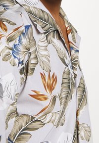 Camicia a maniche corte in tessuto grigio chiaro con grandi stampe di foglie botaniche e fiori in verde, oro, blu e bianco, con colletto morbido.
