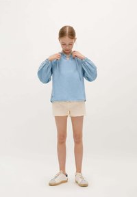 Hellbluse mit langen Ärmeln, einem Schlüssellochausschnitt und Bindedetail, kombiniert mit cremefarbenen Shorts. Texturierte Ärmel mit dekorativen Akzenten.