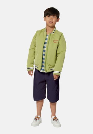Veste bomber verte avec le logo rouge "KENZO PARIS", chemise à carreaux, shorts bleu marine et chaussures blanches avec un détail graphique de tigre.