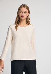 Cremefarbener Langarm-Top mit Bootsausschnitt, versehen mit einer glatten Textur und zwei dekorativen Knöpfen an der Schulter. Kombiniert mit schwarzen Hosen.