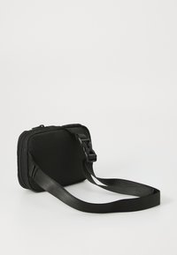 Sac rectangulaire noir avec une bandoulière, vue de côté montrant le matériau texturé et le détail de la fermeture éclair sur un fond blanc.