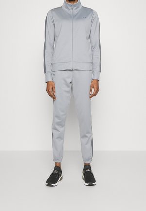 Osoba ubrana w jasnoszary dres z bluzą na zamek i spodniami ze ściągaczami, do tego czarno-białe buty sportowe.