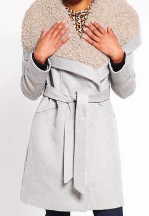 Vrouw die een lichtgrijze jas met riem en grote, gestructureerde kraag over een patroon top aanpast, staand tegen een witte achtergrond.