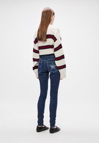 Pullover de malha com listras vermelhas e pretas, combinado com jeans de ganga escuros ajustados com um padrão de estrelas no bolso de trás. Sapatos planos visíveis.