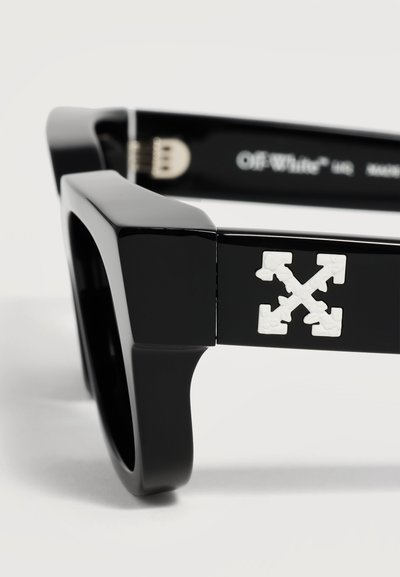 Lunettes de soleil noires à monture épaisse avec un logo blanc à quatre flèches sur la branche et un marquage « Off-White » visible à l'intérieur de la branche.