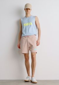 Un débardeur bleu clair avec un texte jaune fluo, associé à un short rose et des baskets blanches. La tenue est complétée par une casquette beige.