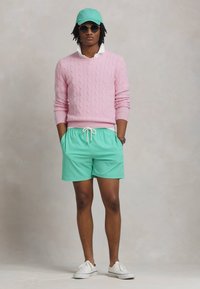 Suéter de malha cabos rosa claro, camisa branca de colarinho, calções verdes menta, boné a condizer e ténis brancos, com óculos de sol como detalhes notáveis.