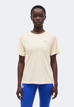 Žena stoji okrenuta prema naprijed, nosi bež Adidas majicu i plave Adidas helanke na jednobojnoj svijetloj pozadini.