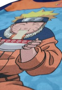 Personnage aux cheveux blonds en pics, portant un bandeau et une veste orange, mangeant des nouilles dans un bol rouge orné de motifs tourbillonnants blancs.