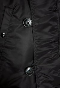 Schwarzer, gepolsterter Jackenstoff mit glatter Textur; verfügt über zwei große, runde schwarze Knöpfe und sichtbare Nähte.