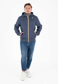Sweat à capuche zippé marine avec une fermeture éclair jaune et blanche, poches kangourou et capuche avec cordon, associé à un jean bleu et des baskets blanches.