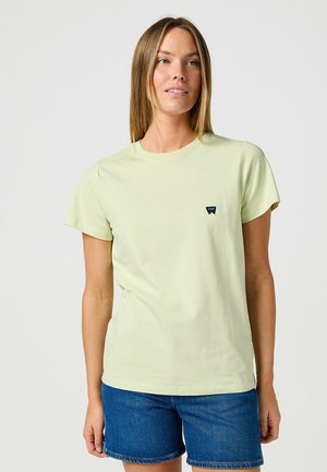 Hellgrünes Baumwoll-T-Shirt mit rundem Halsausschnitt und kurzen Ärmeln. Verfügt über ein kleines dunkelgraues Logo auf der linken Brust. Kombiniert mit blauen Jeansshorts.