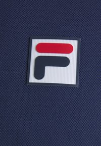 Écusson du logo FILA avec des formes circulaires rouges et bleu foncé sur un fond blanc texturé, fixé sur une surface en tissu bleu marine.