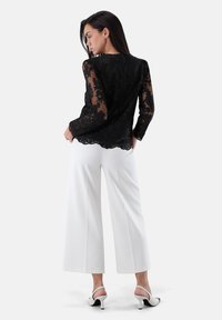 Schwarzes Spitzentop mit transparenten langen Ärmeln, gezacktem Saum; kombiniert mit weißen, weit geschnittenen Hosen und weißen Sandaletten mit Absatz.