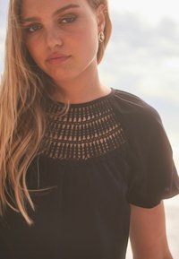 Sort top med en detaljeret hæklet krave. Stoffet ser blødt ud, med en løs pasform og korte ærmer. Gyldne hoops-øreringe fremhæver looket.