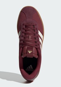 Bordové semišové tenisky s bílými pruhy, gumovou podrážkou a zlatými akcenty. Mají polstrovaný límec a šněrovací design.