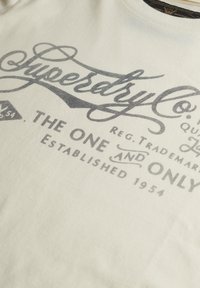 Tissu blanc avec texte gris imprimé "Superdry Co. The One and Only Established 1954" en police stylisée sur un t-shirt.