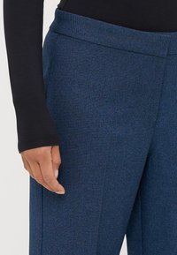 Blauwe textuurbroek met een platte tailleband. Zichtbare zijzak. De stof lijkt zacht met een subtiel weefpatroon. Hand rustend op de stof.