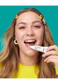 Baume à lèvres Ceramidin dans un tube blanc à presser, tenu par une personne portant un haut jaune vif et des accessoires capillaires, sur un fond teal.