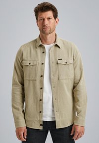 Chemise beige à boutons avec un tissage texturé, deux poches poitrine et des boutons foncés, superposée à un t-shirt blanc. Portée avec un jeans noir.