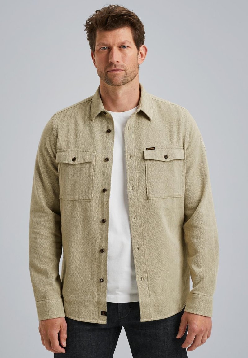 Camicia beige con bottoni, a tessuto intrecciato, due tasche sul petto e bottoni scuri, indossata sopra una maglietta bianca. Abbinata a jeans scuri.
