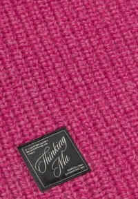 Gros plan sur un tissu tricoté texturé rose avec une étiquette rectangulaire noire portant la mention "Thinking Mu" et "Une touche méditerranéenne contemporaine."