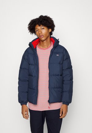 Giubbino imbottito blu navy con accenti rossi. Presenta un cappuccio, chiusura a zip e polsini elasticizzati. Indossato sopra una maglietta a righe rossa e bianca.
