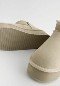 Beżowe zamszowe buty na platformie mają okrągły czubek, wysokość do kostki, zamek błyskawiczny z boku oraz teksturowaną gumową podeszwę z pionowymi rowkami.