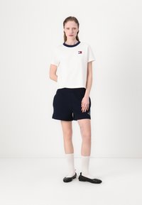 Hvid bomulds T-shirt med marineblå krave og lille logo, parret med marineblå shorts med elastik i taljen. Modellen bærer hvide sokker og sorte flade sko.