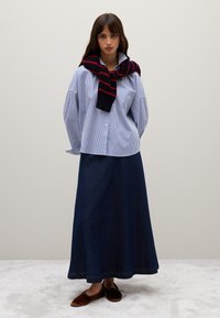 Gestreifte Button-Down-Bluse mit hochgekrempelten Ärmeln, kombiniert mit einem langen Jeansrock. Ein schwarz-roter gestreifter Pullover um den Hals gebunden. Braune Slipper.