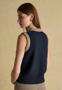 Navy blue sleeveless sweater with a beige collar detail, made of soft material. Textured fabric and a relaxed fit create a casual look.
