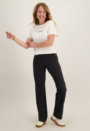 Lachende vrouw met lang blond haar draagt een wit "Couture" T-shirt, zwarte broek en witte sneakers, staand tegen een effen achtergrond.