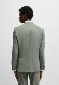 Veste de costume gris clair en tissu texturé. Elle présente un col cranté, une fente simple au dos et des boutons argentés aux poignets.