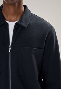 Tmavomodrá bunda s predným zipsom, textúrovaný materiál, golierový dizajn s predným vreckom, strieborný zips, minimalistický štýl.