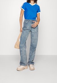 Blå texturerad kortärmad topp med vita detaljer, tillsammans med urtvättade blå denimjeans med blommiga och texttryck, samt beige klumpiga skor.