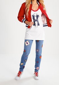 Czerwona kurtka bombka z białymi i granatowymi akcentami na białym topie z granatowym logo. Jasnoniebieskie dżinsy z kolorowymi łatkami. Białe sneakersy.