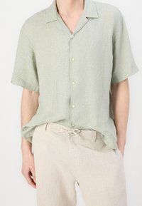 Světlezelená košile s krátkým rukávem z lněné látky, s límečkem a zapínáním na knoflíky. V kombinaci s béžovými kalhotami na šňůrku.