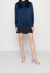 Kvinde iført en marineblå satinbluse, sort plisseret miniskirt og lyse lilla blokhælede sandaler, der står mod en ensfarvet baggrund.