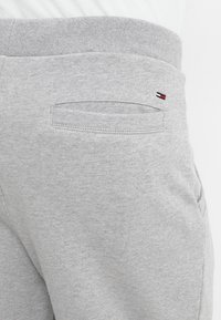 Zbliżenie na kieszeń tylnią jasnoszarych spodni dresowych z małym prostokątnym logo w kolorach czerwonym, białym i czarnym nad kieszenią na pasku.