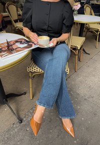 Melnā puff-enģeļa piedurkņu blūze, vidēji zilās platās bikses un garas, glancētas brūnas kurpes ar norādītu purngalu. Rokā tur tasi uz šķīvja, blakus atrodas žurnāls.