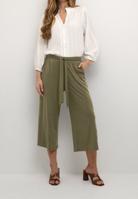 Vit blus med långa ärmar och spetsdetaljer, kombinerad med olivgröna byxor med vida ben och knytliv. Bruna klacksandaler fulländar looken.