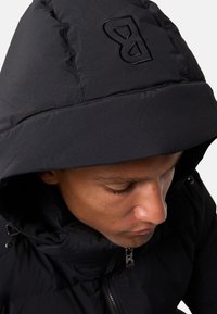 Zwarte pufferjack met een grote capuchon en een rits aan de voorkant. Bevat een reliëflogo op de capuchon en een textuurstof met een matte afwerking.