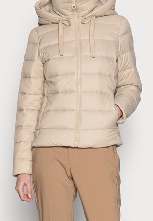 Doudoune matelassée beige avec capuche et fermeture éclair à l'avant, portée avec un pantalon marron clair, main dans la poche latérale, sur fond uni.